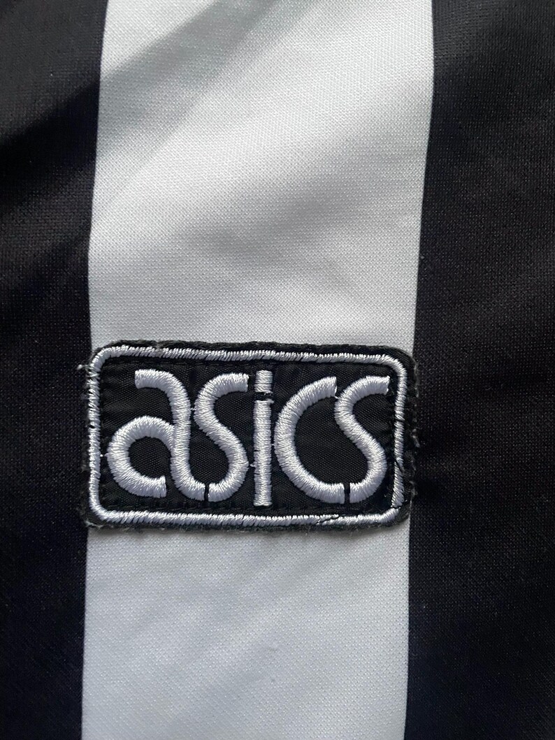 May include: Close-up of a black and white striped fabric with an embroidered ASICS logo patch. The logo is white on a black background, with a white border. The fabric has vertical black and white stripes.