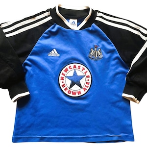 K&ouml;nnte beinhalten: Ein blau-schwarzes Langarm-Fu&szlig;balltrikot mit dem Newcastle United-Wappen und einem Sternlogo. Das Trikot hat wei&szlig;e Streifen auf den &Auml;rmeln und das Adidas-Logo.