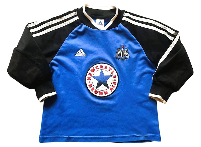 K&ouml;nnte beinhalten: Ein blau-schwarzes Langarm-Fu&szlig;balltrikot mit dem Newcastle United-Wappen und einem Sternlogo. Das Trikot hat wei&szlig;e Streifen auf den &Auml;rmeln und das Adidas-Logo.