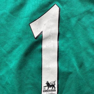 K&ouml;nnte beinhalten: Nahaufnahme eines gr&uuml;nen Stoffes mit einer gro&szlig;en wei&szlig;en Zahl 1. Die Zahl hat eine schwarze Umrandung und ein abgenutztes Aussehen. Das Logo der F.A. Premier League ist unten.