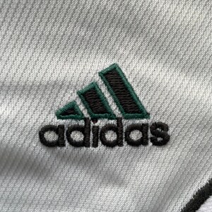Puede incluir: Primer plano de una tela blanca con el logotipo de Adidas bordado en negro y verde. El logotipo presenta tres rayas verdes sobre el nombre de la marca.