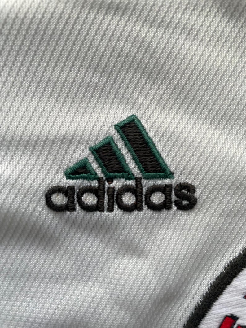 Puede incluir: Primer plano de una tela blanca con el logotipo de Adidas bordado en negro y verde. El logotipo presenta tres rayas verdes sobre el nombre de la marca.