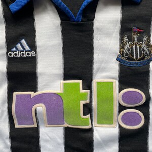 May include: Black and white striped football shirt with a blue collar and the Adidas logo on the left breast. The shirt has a green and purple "ntl" logo on the front and the Newcastle United crest on the right breast.