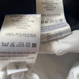 Pu&ograve; includere: Etichetta di cura per abbigliamento bianca e nera con testo in tedesco, inglese e francese. L'etichetta indica "100% Poliestere" e include istruzioni per il lavaggio e la stiratura. Il testo recita "Die Weltmarke mit den 3 Streifen", "The Brand with the 3 Stripes" e "La Marque aux 3 Bandes".