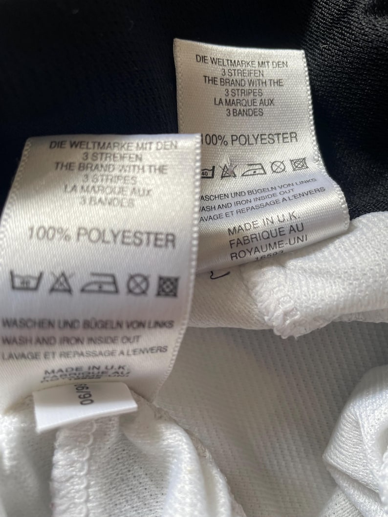 Pu&ograve; includere: Etichetta di cura per abbigliamento bianca e nera con testo in tedesco, inglese e francese. L'etichetta indica "100% Poliestere" e include istruzioni per il lavaggio e la stiratura. Il testo recita "Die Weltmarke mit den 3 Streifen", "The Brand with the 3 Stripes" e "La Marque aux 3 Bandes".