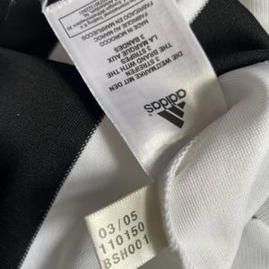 May include: Close-up of a white garment with black stripes. The Adidas logo is visible, along with tags displaying text in multiple languages, including "3 STREIFEN" and "3 STRIPES". The garment's fabric appears to be a smooth, possibly athletic material.