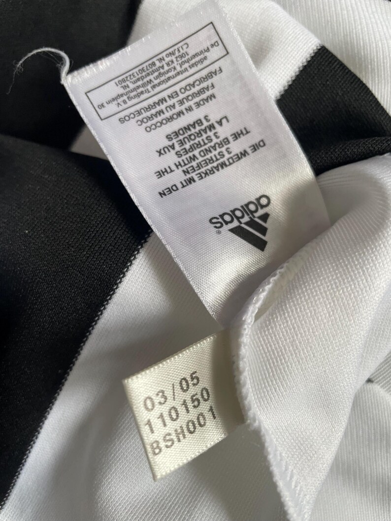 May include: Close-up of a white garment with black stripes. The Adidas logo is visible, along with tags displaying text in multiple languages, including "3 STREIFEN" and "3 STRIPES". The garment's fabric appears to be a smooth, possibly athletic material.