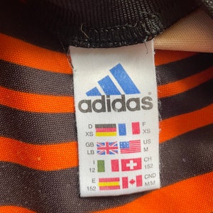 May include: A white tag with the Adidas logo and size chart. The tag is black and white with the Adidas logo in blue. The size chart includes sizes for Germany, France, United Kingdom, United States, Switzerland, and Canada. The sizes are XS, M, 152, and M/M.