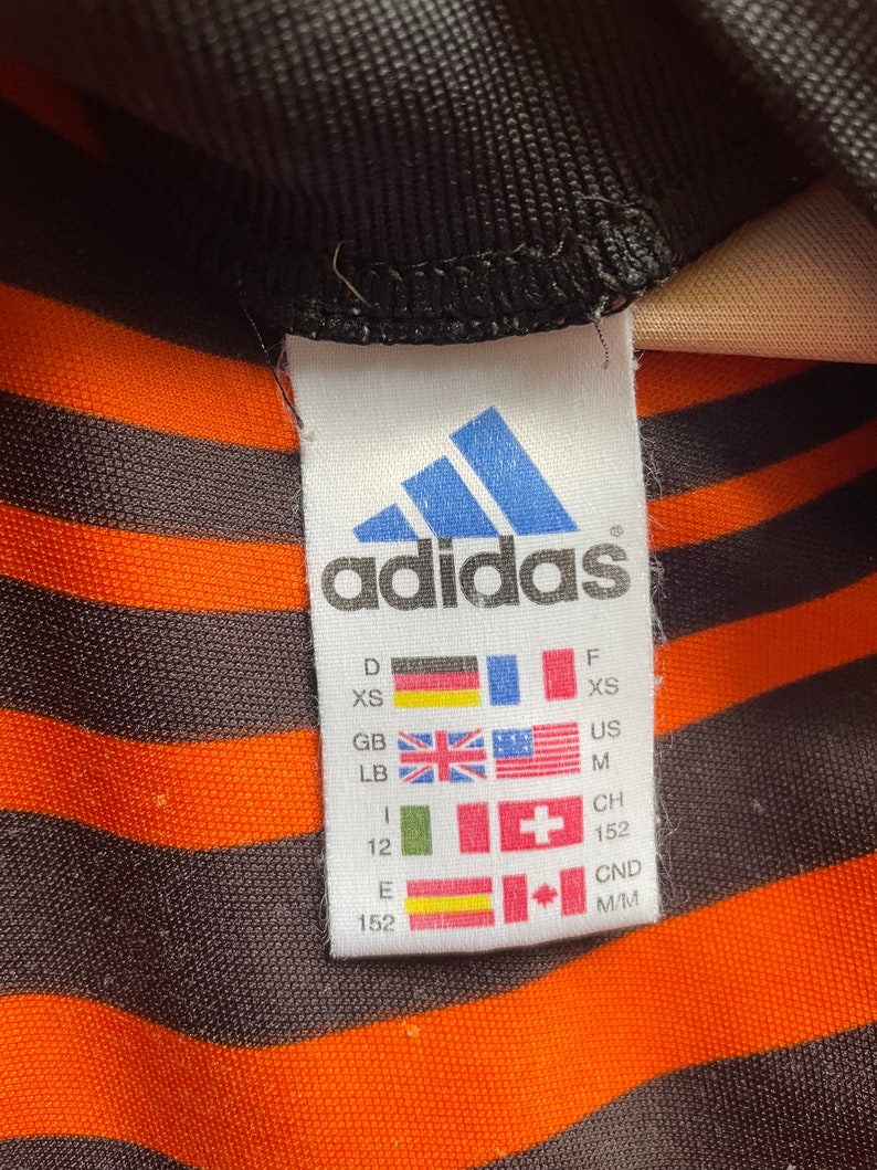 May include: A white tag with the Adidas logo and size chart. The tag is black and white with the Adidas logo in blue. The size chart includes sizes for Germany, France, United Kingdom, United States, Switzerland, and Canada. The sizes are XS, M, 152, and M/M.