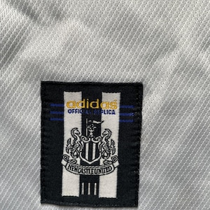 May include: A white soccer jersey with a black and white adidas logo and a black and white Newcastle United crest. The logo says "adidas Official Replica".