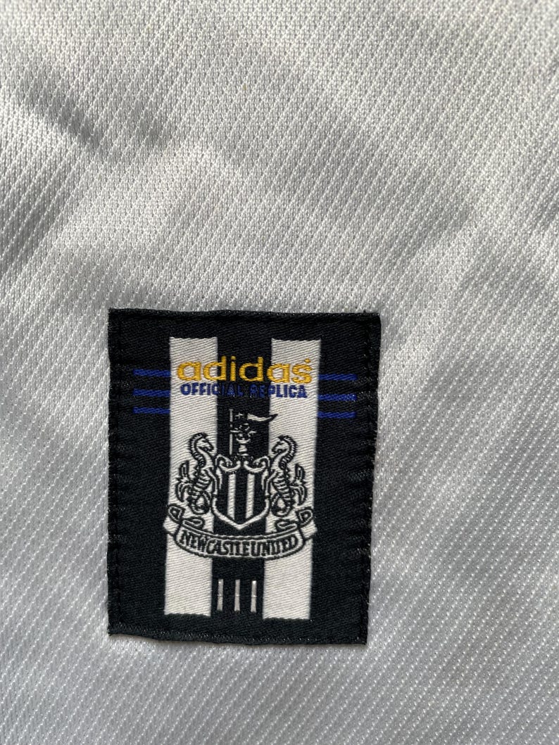 May include: A white soccer jersey with a black and white adidas logo and a black and white Newcastle United crest. The logo says "adidas Official Replica".