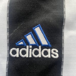 May include: Close-up of a black and white striped fabric with the Adidas logo embroidered in white with blue accents. The Adidas logo is composed of three slanted stripes and the brand name.