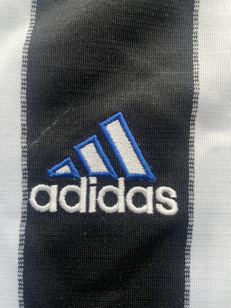 May include: Close-up of a black and white striped fabric with the Adidas logo embroidered in white with blue accents. The Adidas logo is composed of three slanted stripes and the brand name.