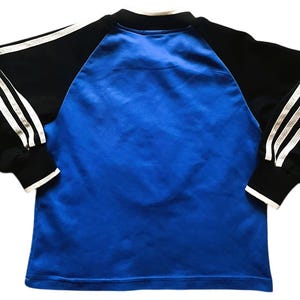 K&ouml;nnte beinhalten: Ein blau-schwarzes Langarmshirt. Das Shirt hat einen blauen K&ouml;rper mit schwarzen &Auml;rmeln. Die &Auml;rmel haben drei wei&szlig;e Streifen entlang der Arml&auml;nge. Die Manschetten und der Kragen sind wei&szlig; eingefasst.