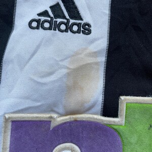 May include: A white and black soccer jersey with the Adidas logo and the number 6 on the front. The jersey has a green and purple trim around the number.