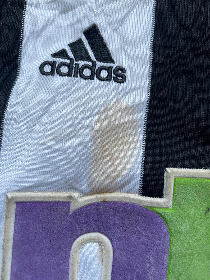 May include: A white and black soccer jersey with the Adidas logo and the number 6 on the front. The jersey has a green and purple trim around the number.
