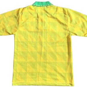 K&ouml;nnte beinhalten: Ein gelbes Fu&szlig;balltrikot mit kurzen &Auml;rmeln, gr&uuml;nem Kragen und einem geometrischen Muster aus d&uuml;nnen gr&uuml;nen Linien.