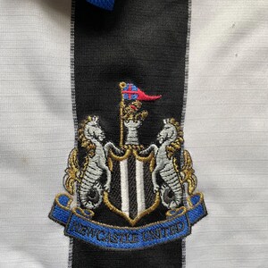 May include: A black and white striped soccer jersey with a blue collar. The jersey features an embroidered crest with a black and white shield, two gold seahorses, and the text "NEWCASTLE UNITED" in a blue ribbon.