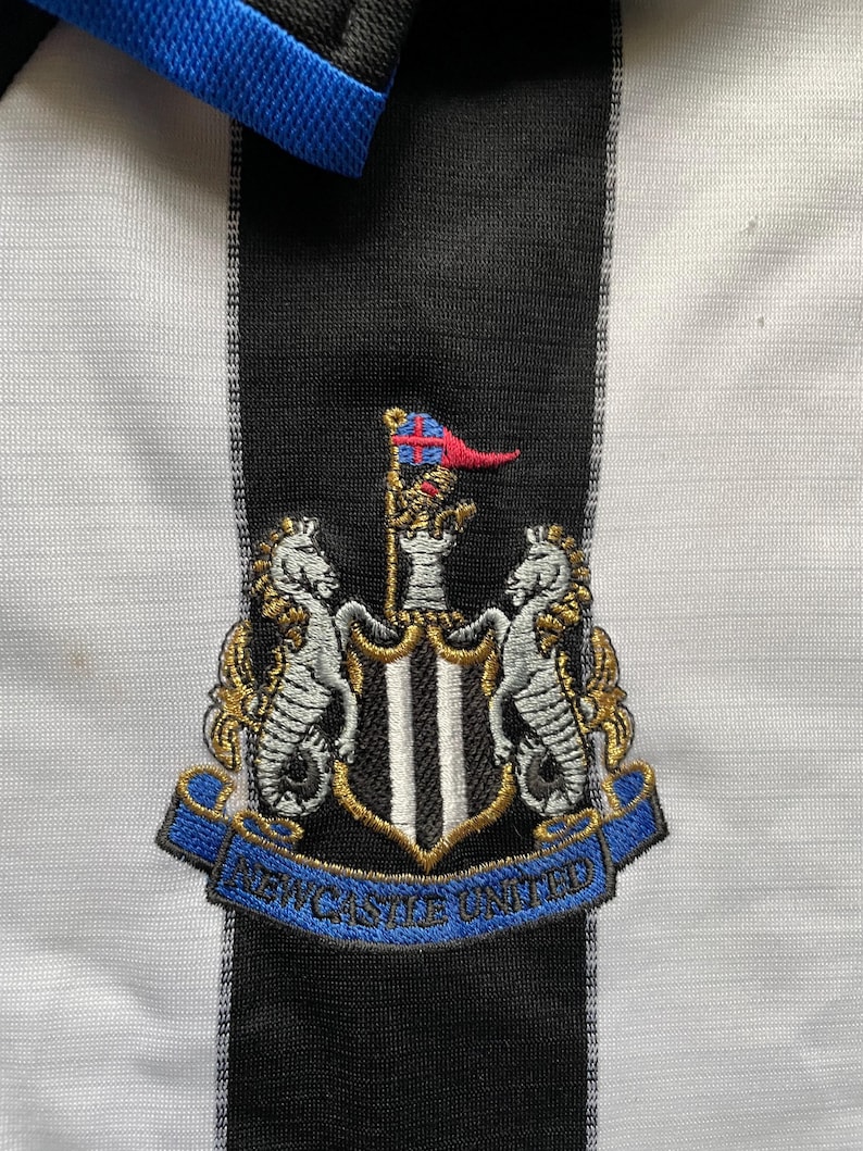 May include: A black and white striped soccer jersey with a blue collar. The jersey features an embroidered crest with a black and white shield, two gold seahorses, and the text "NEWCASTLE UNITED" in a blue ribbon.