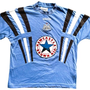 May include: A light blue t-shirt with black and white stripes on the sleeves. The shirt has a large Newcastle Brown Ale logo on the front with a star and the words "Newcastle Brown Ale". The shirt also has the Adidas logo on the front.