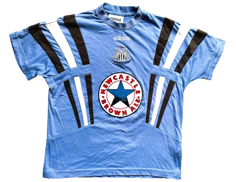 May include: A light blue t-shirt with black and white stripes on the sleeves. The shirt has a large Newcastle Brown Ale logo on the front with a star and the words "Newcastle Brown Ale". The shirt also has the Adidas logo on the front.