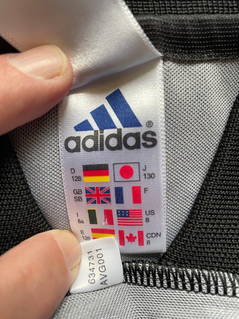 May include: White tag with black adidas logo and size chart in various languages and measurements. The tag also includes the numbers 634731 and AVG001.