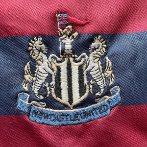 May include: A close-up of a red and navy blue striped shirt. The shirt displays an embroidered crest with two seahorses, a shield, and the words "Newcastle United" in a blue banner.