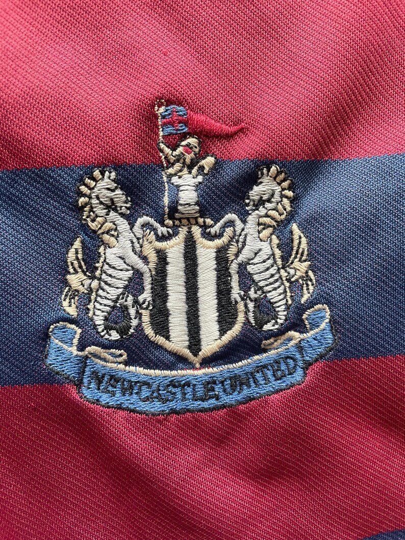 May include: A close-up of a red and navy blue striped shirt. The shirt displays an embroidered crest with two seahorses, a shield, and the words "Newcastle United" in a blue banner.