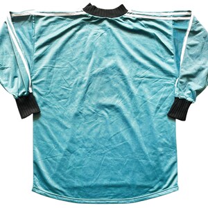 May include: A long-sleeved teal jersey with white stripes on the shoulders and sleeves. The cuffs and collar are black. The jersey is made of a soft, textured material.