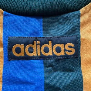 May include: Close-up of a vintage Adidas athletic jersey. The jersey features a color block design with blue, green, and gold vertical stripes. The Adidas logo is prominently displayed in gold lettering on a black patch.