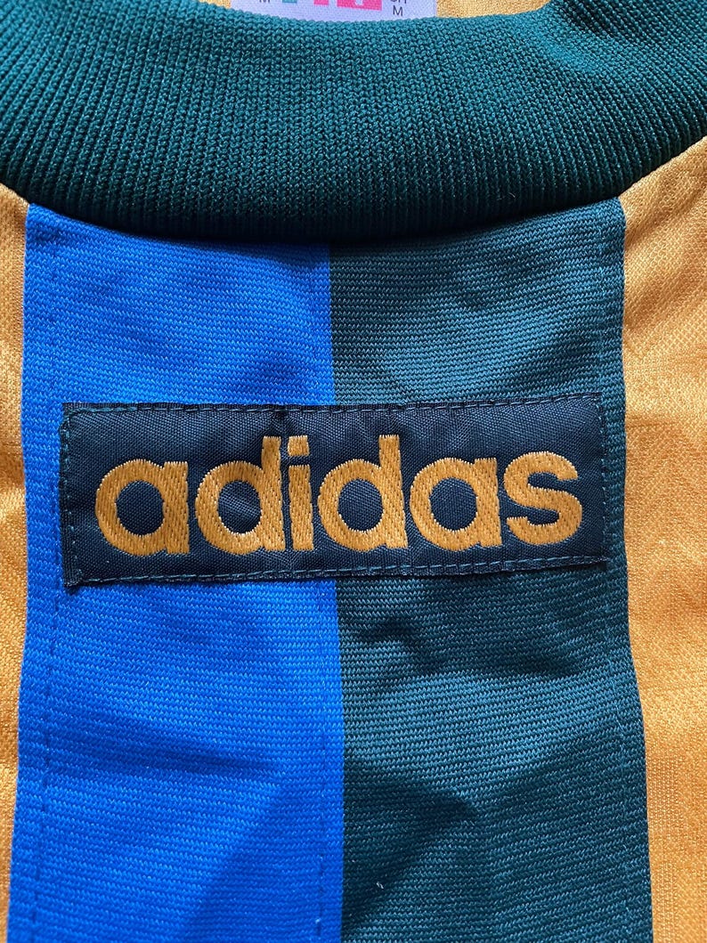 May include: Close-up of a vintage Adidas athletic jersey. The jersey features a color block design with blue, green, and gold vertical stripes. The Adidas logo is prominently displayed in gold lettering on a black patch.
