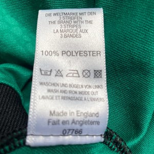 K&ouml;nnte beinhalten: Nahaufnahme eines wei&szlig;en Etiketts mit Text in mehreren Sprachen, darunter "DIE WELTMARKE MIT DEN 3 STREIFEN" und "100% POLYESTER". Das Etikett ist an einem gr&uuml;n-schwarzen Stoff befestigt. Das Etikett sagt auch "Made in England".