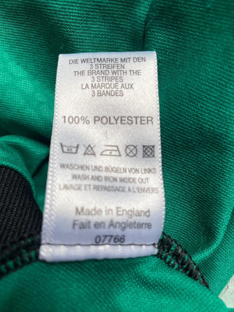 K&ouml;nnte beinhalten: Nahaufnahme eines wei&szlig;en Etiketts mit Text in mehreren Sprachen, darunter "DIE WELTMARKE MIT DEN 3 STREIFEN" und "100% POLYESTER". Das Etikett ist an einem gr&uuml;n-schwarzen Stoff befestigt. Das Etikett sagt auch "Made in England".