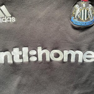 May include: Black football shirt with a white Adidas logo, white "ntl:home" text, and a white and black football team crest featuring a black and white bird.