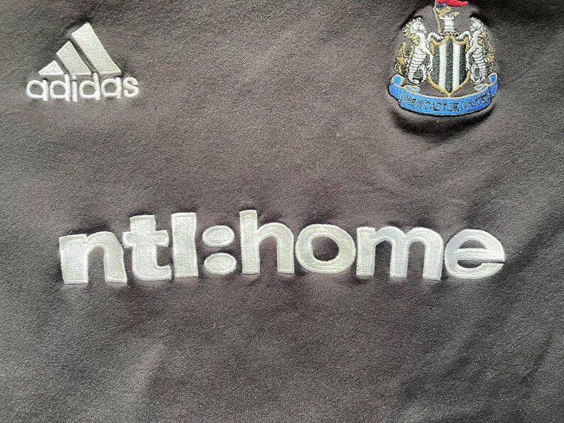 May include: Black football shirt with a white Adidas logo, white "ntl:home" text, and a white and black football team crest featuring a black and white bird.
