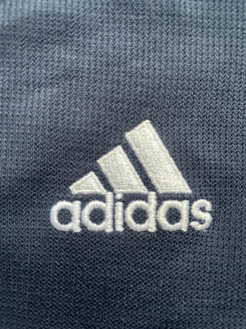 Puede incluir: Primer plano de una tela azul marino con un logotipo de Adidas bordado en blanco.