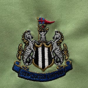 Pu&ograve; includere: Primo piano di un tessuto verde con uno stemma ricamato. Lo stemma presenta due cavallucci marini che fiancheggiano uno scudo a strisce bianche e nere. Uno stendardo blu recita "NEWCASTLE UNITED". Lo stemma &egrave; dettagliato con accenti oro, bianchi e rossi.