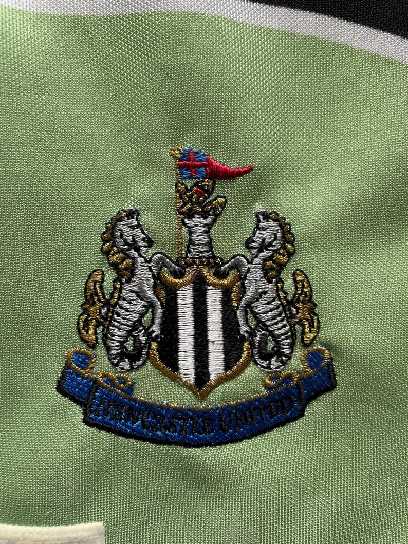 Pu&ograve; includere: Primo piano di un tessuto verde con uno stemma ricamato. Lo stemma presenta due cavallucci marini che fiancheggiano uno scudo a strisce bianche e nere. Uno stendardo blu recita "NEWCASTLE UNITED". Lo stemma &egrave; dettagliato con accenti oro, bianchi e rossi.