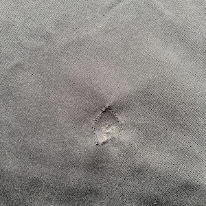 May include: Close-up of a dark gray fabric with a visible tear. The fabric has a textured appearance, and the tear is located near the center of the image. The edges of the tear are frayed, revealing the inner layers of the material.