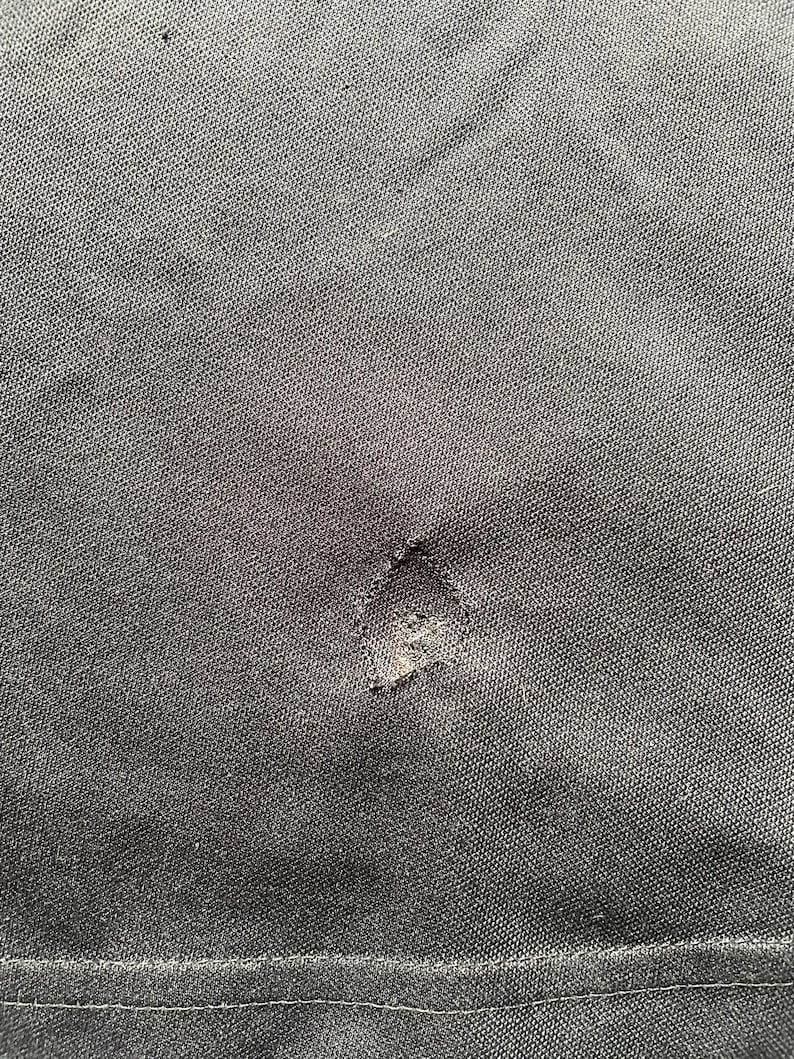 May include: Close-up of a dark gray fabric with a visible tear. The fabric has a textured appearance, and the tear is located near the center of the image. The edges of the tear are frayed, revealing the inner layers of the material.
