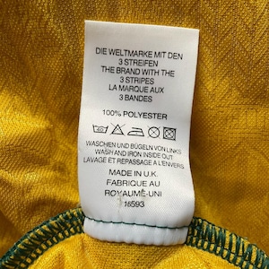 May include: Close-up of a tag on a yellow and green garment. The tag reads "THE BRAND WITH THE 3 STRIPES" and "100% POLYESTER." The garment is made in the U.K. and has a green stitched hem.