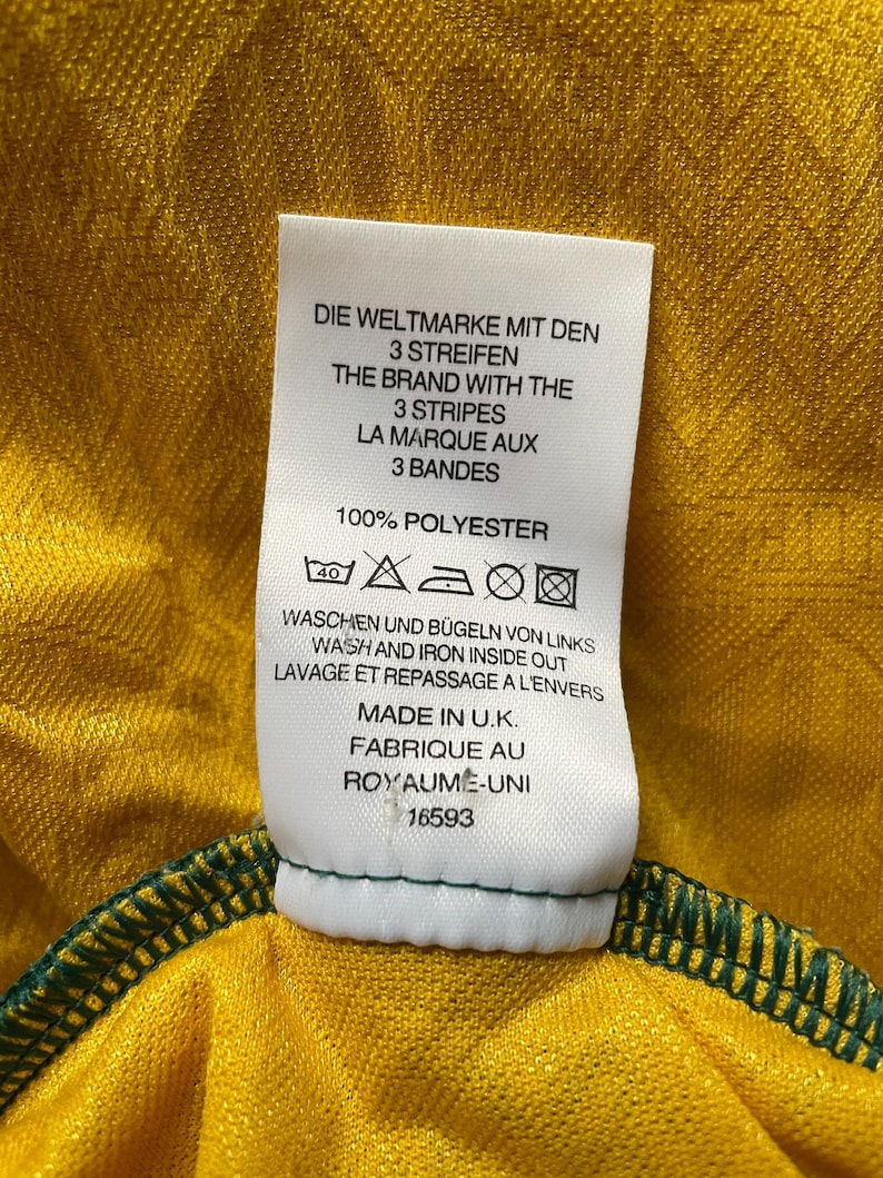 May include: Close-up of a tag on a yellow and green garment. The tag reads "THE BRAND WITH THE 3 STRIPES" and "100% POLYESTER." The garment is made in the U.K. and has a green stitched hem.
