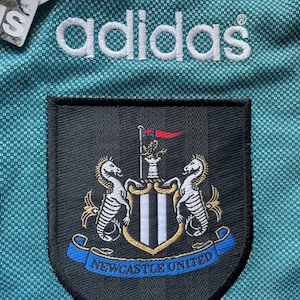 K&ouml;nnte beinhalten: Nahaufnahme eines t&uuml;rkisgr&uuml;nen Fu&szlig;balltrikots mit dem Adidas-Logo in Wei&szlig;. Das Wappen von Newcastle United ist zu sehen, mit zwei Seepferdchen und dem Schriftzug "Newcastle United" auf einem blauen Banner.