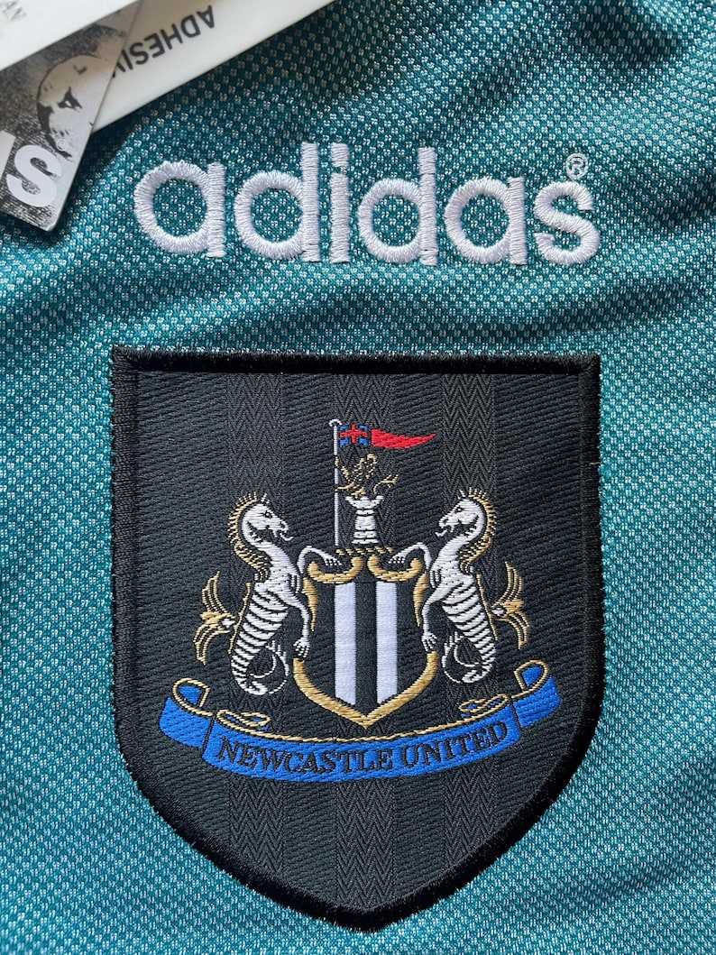 K&ouml;nnte beinhalten: Nahaufnahme eines t&uuml;rkisgr&uuml;nen Fu&szlig;balltrikots mit dem Adidas-Logo in Wei&szlig;. Das Wappen von Newcastle United ist zu sehen, mit zwei Seepferdchen und dem Schriftzug "Newcastle United" auf einem blauen Banner.