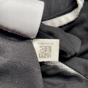 Pu&ograve; includere: Primo piano di una maglia sportiva nera con finiture bianche e un'etichetta. L'etichetta mostra il testo "KDSMD6H201287" e un codice QR. La maglia ha un motivo a righe strutturato.