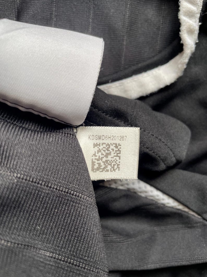 Pu&ograve; includere: Primo piano di una maglia sportiva nera con finiture bianche e un'etichetta. L'etichetta mostra il testo "KDSMD6H201287" e un codice QR. La maglia ha un motivo a righe strutturato.