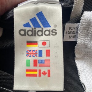 May include: A white tag with black text that reads "adidas" and a blue adidas logo. The tag also has a black and white label with the text "907417 ASR001/12/02". The tag has a variety of flags printed on it, including the flags of Germany, Japan, the United Kingdom, France, Italy, the United States, Spain, and Canada.