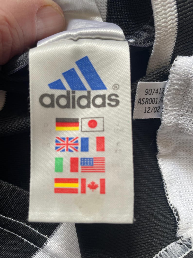 May include: A white tag with black text that reads "adidas" and a blue adidas logo. The tag also has a black and white label with the text "907417 ASR001/12/02". The tag has a variety of flags printed on it, including the flags of Germany, Japan, the United Kingdom, France, Italy, the United States, Spain, and Canada.