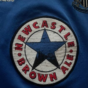 K&ouml;nnte beinhalten: Nahaufnahme eines blauen Fu&szlig;balltrikots mit dem Adidas-Logo und einem Newcastle Brown Ale-Patch. Der Patch ist kreisf&ouml;rmig mit einem blauen Stern und den Worten "Newcastle Brown Ale" in Rot.