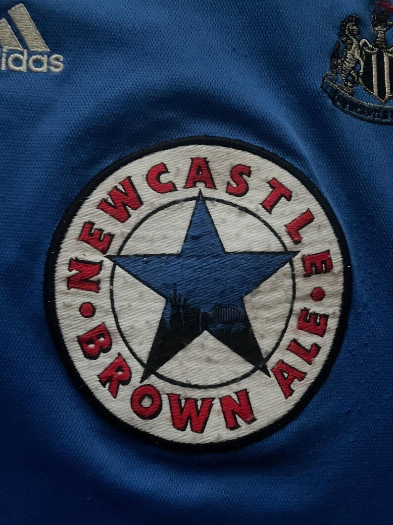 K&ouml;nnte beinhalten: Nahaufnahme eines blauen Fu&szlig;balltrikots mit dem Adidas-Logo und einem Newcastle Brown Ale-Patch. Der Patch ist kreisf&ouml;rmig mit einem blauen Stern und den Worten "Newcastle Brown Ale" in Rot.